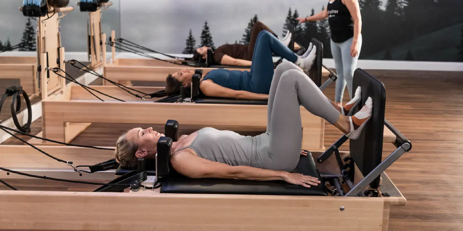 Instrutora orienta uma aluna durante uma aula de grupo de pilates em estúdio, com equipamentos como reformer; pessoas praticam alongamento e postura em grupo.