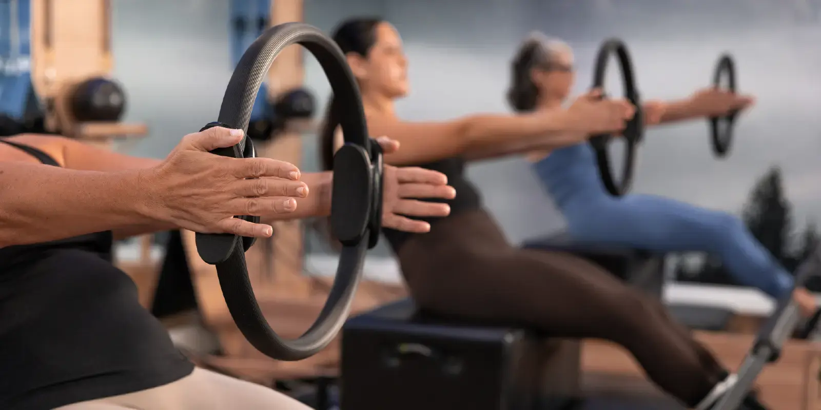 Grupo de alunas de Pilates sentadas na box, em cima do reformer. Exercício de Pilates de braços esticados a pressionar o Magic Circle para trabalhar o core.