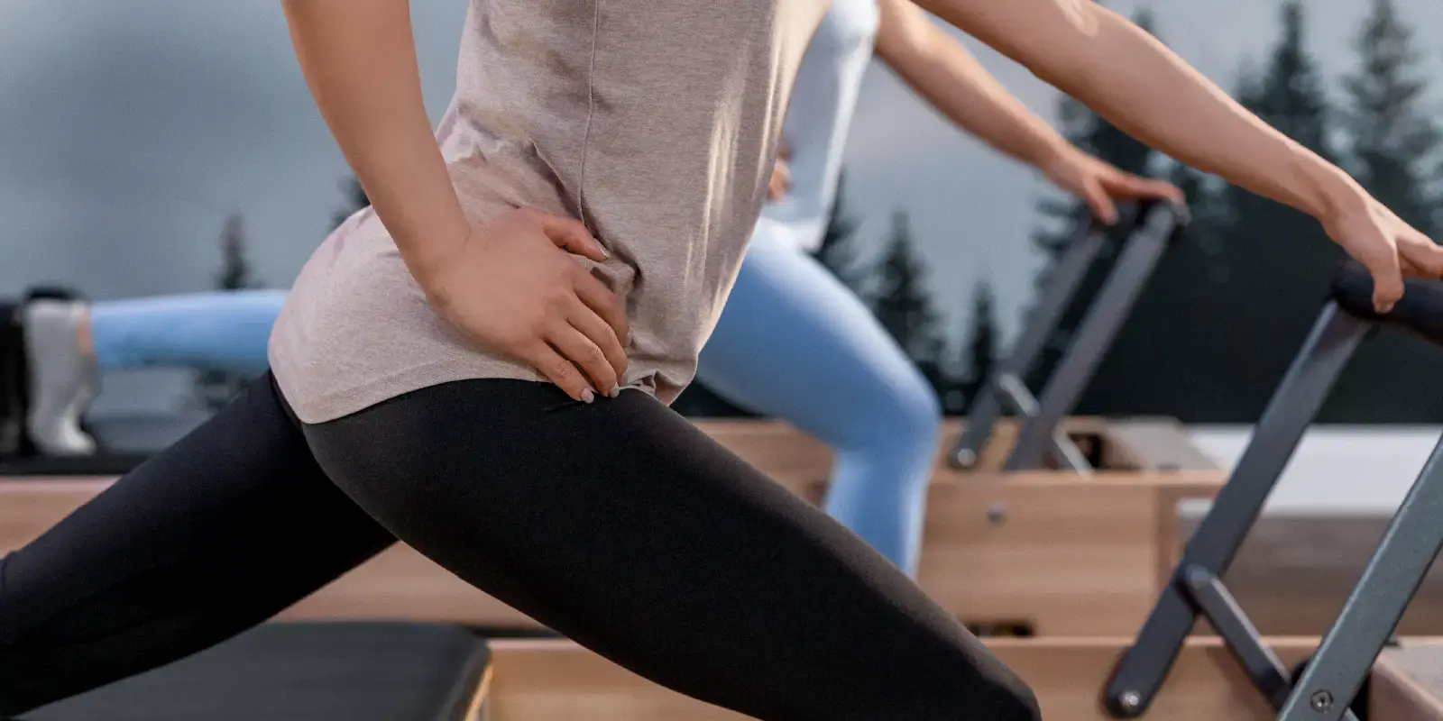Grupo de mulheres a treinar no reformer durante uma aula de grupo de Pilates. As mulheres estão de pé ao lado do reformer com um pé apoiado no chão e outro pé apoiado na pega do reformer. Uma mão está apoiada na anca e a outra mão está apoiada na footbar. O grupo está a realizar o exercício Single Leg Front Split. 